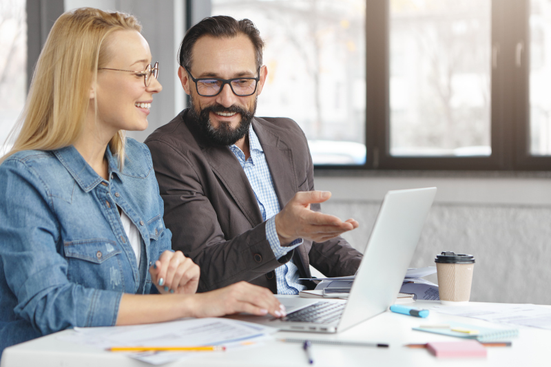 Experienced mature male manager expalins business strategy to young colleague, shows information on laptop computer has pleased expression, glad to work and collaborate together. Two corporate workers