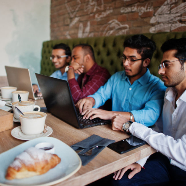 Group of four south asian men's posed at business meeting in cafe. Indians work with laptops together using various gadgets, having conversation.